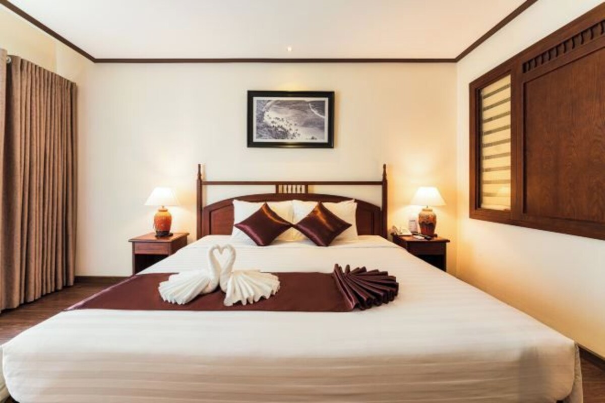 Een netjes opgemaakt bed met een bruine plaid, twee kussens, en een decoratieve zwaan en waaier compositie. Er staan twee nachtlampjes en een ingelijst schilderij aan de muur boven het bed.