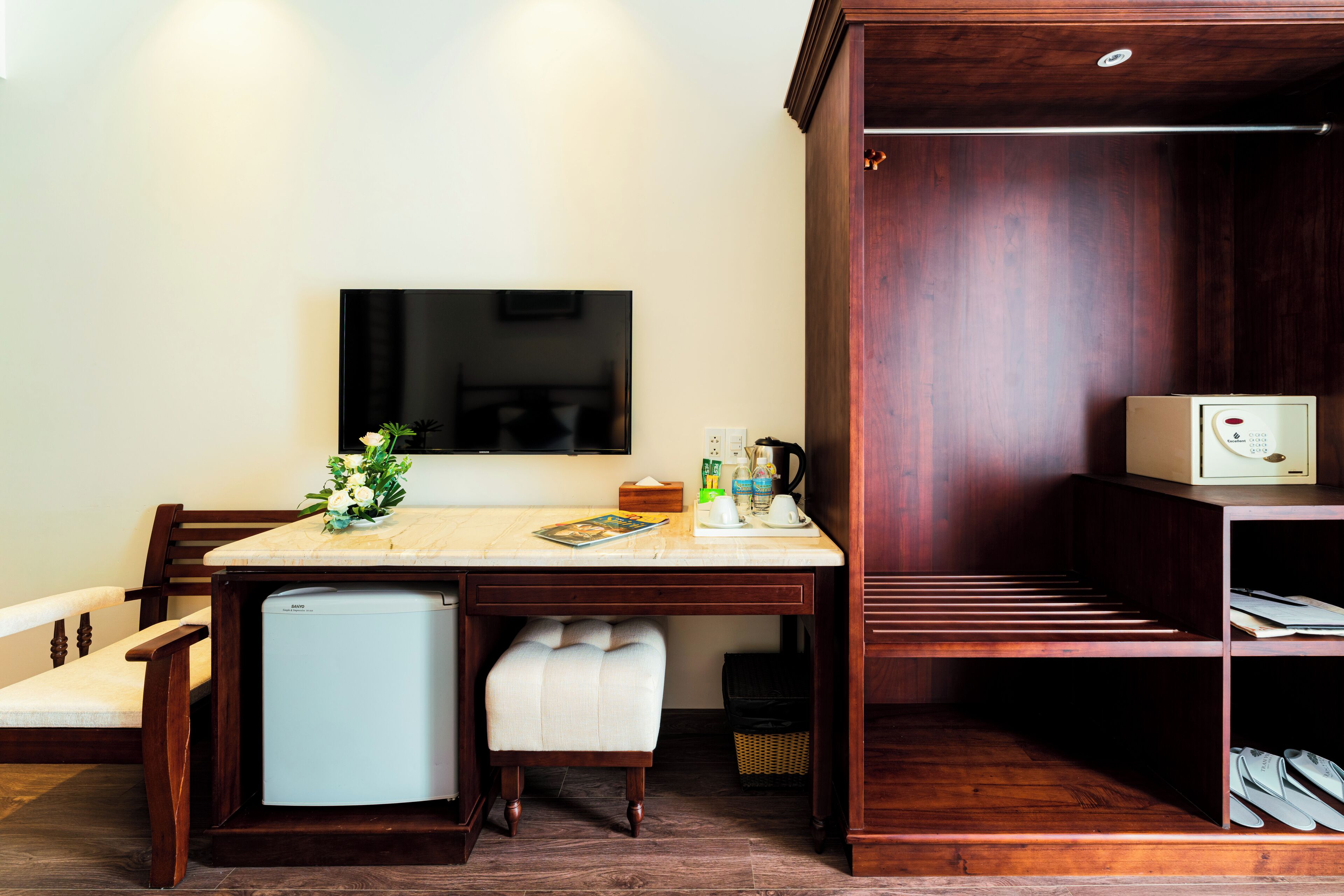 Une chambre d’hôtel compacte comprenant un bureau en bois, un petit réfrigérateur, une télévision, une armoire en bois et un porte-chaussures.