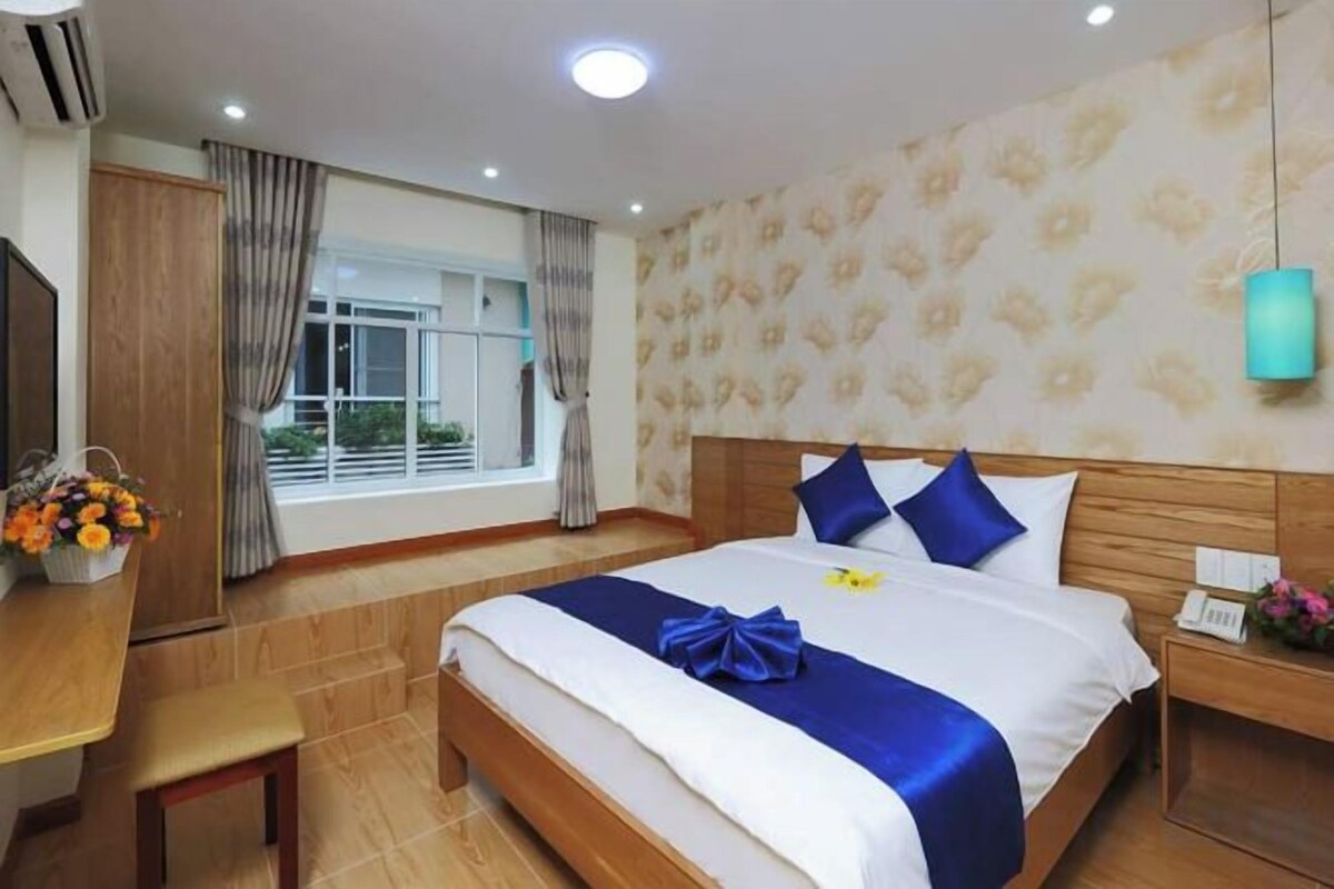 A hotel room with a large bed, a desk, a chair, a window with curtains, and a wall-mounted lamp.
