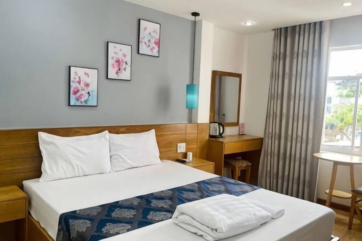 A hotel room with a bed, bedside tables, a desk, a chair, a lamp, a mirror, and a window with curtains.