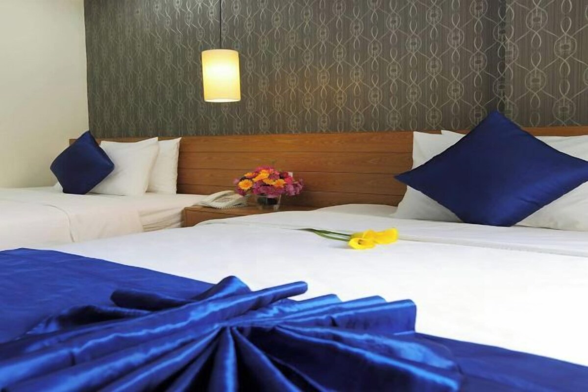 A hotel room with two beds, a wooden headboard, a nightstand with a lamp, and a vase with flowers.