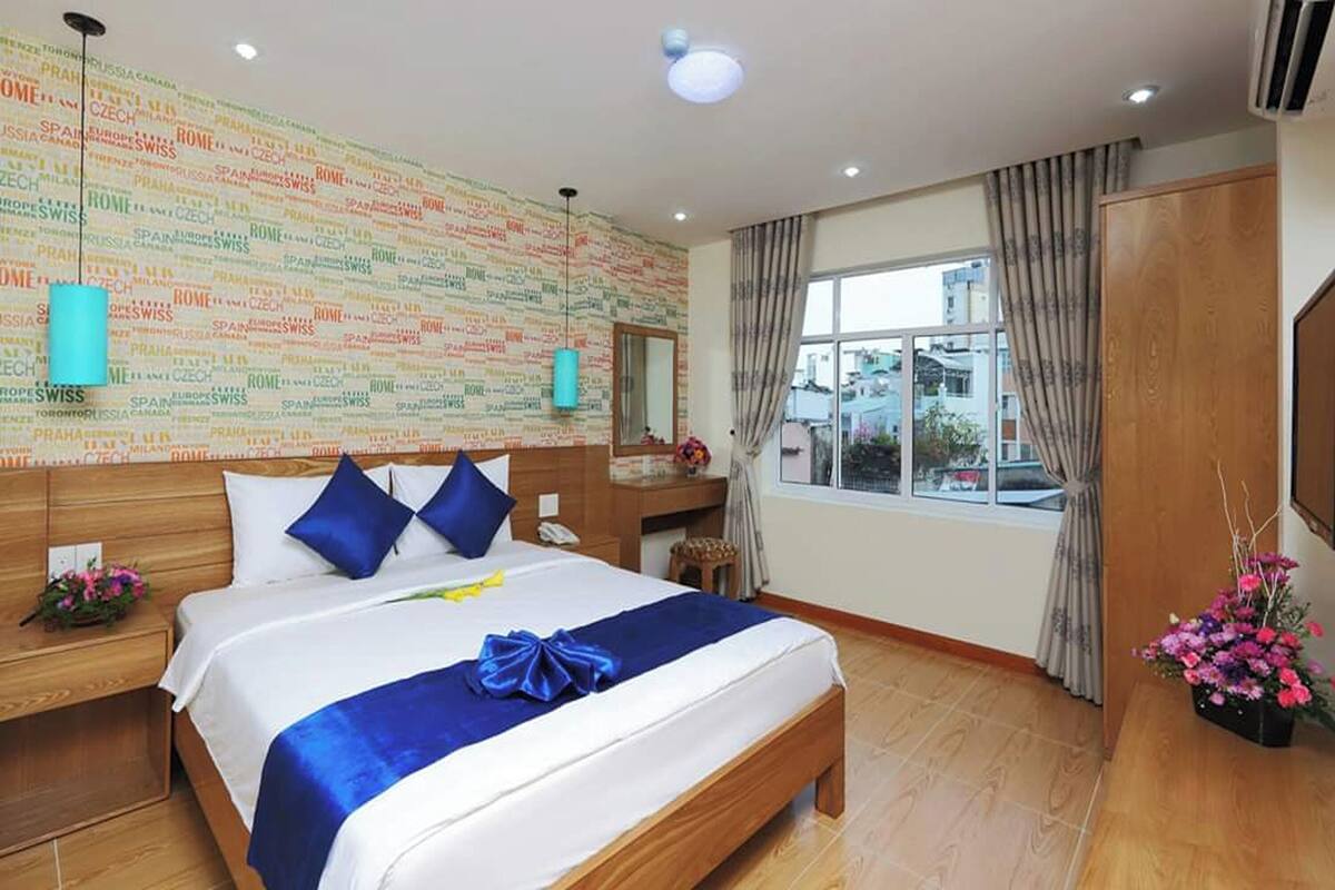 A hotel room with a large bed, a wooden headboard, a wall with international city names, a window with curtains, and a wooden wardrobe.