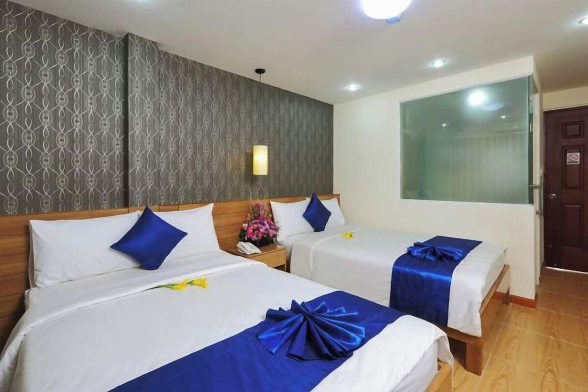 A hotel room with two beds, each with white linens and blue accents, a wooden headboard, a bedside table with a lamp, and a large window.