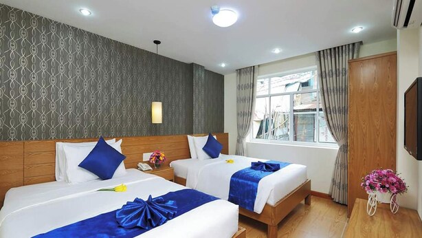 A hotel room with two beds, a wooden headboard, a window with curtains, a wall-mounted TV, and a small table with a flower arrangement. - Ailen Hotel (Ho Chi Minh City)