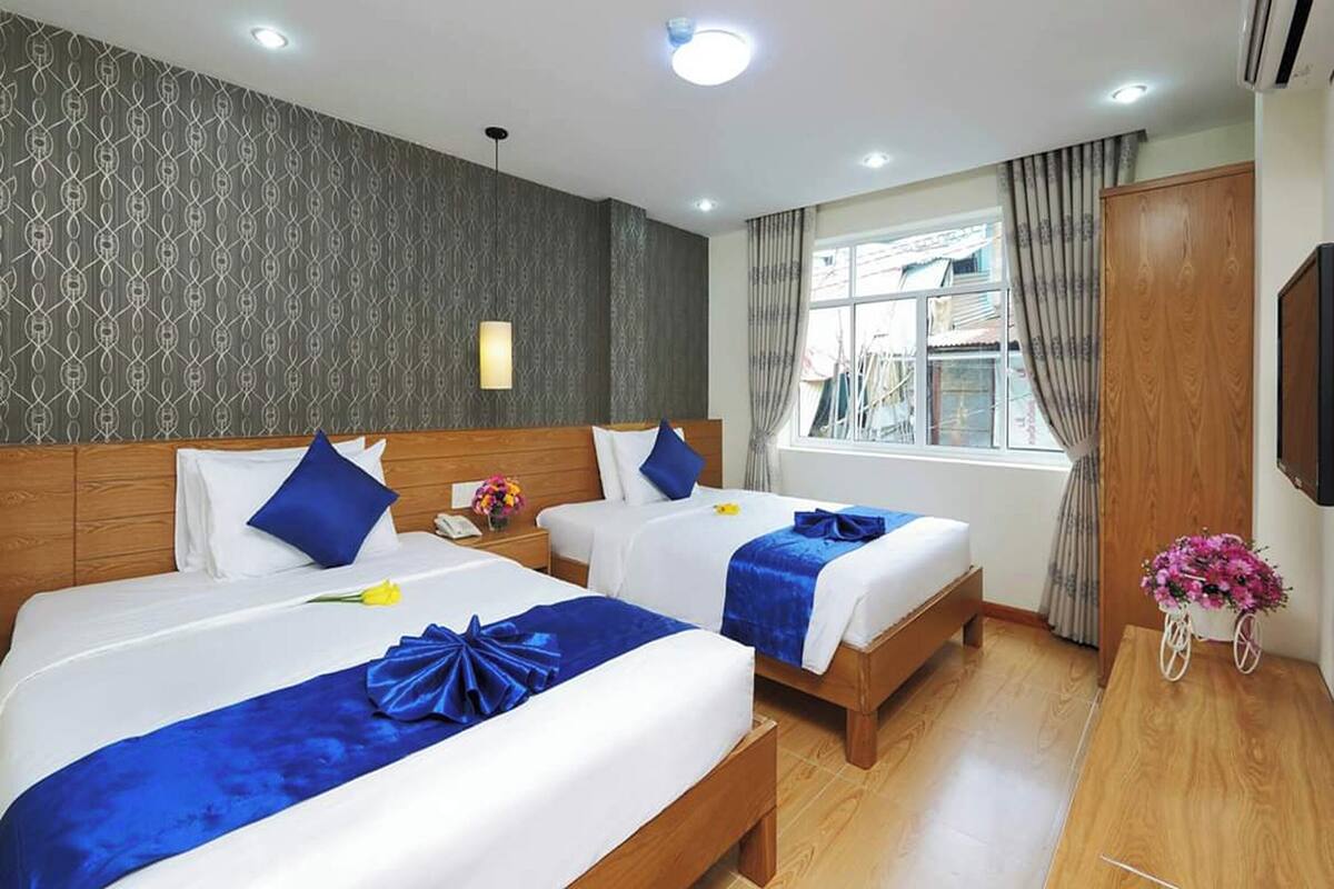 A hotel room with two beds, a wooden headboard, a window with curtains, a wall-mounted TV, and a small table with a flower arrangement.