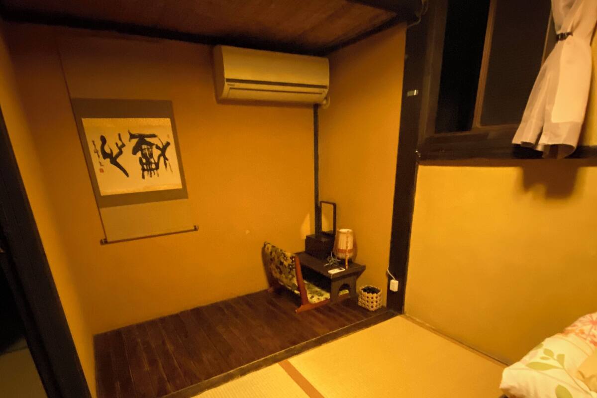 A traditional Japanese-style room with tatami flooring, a futon bed, a small bedside table with a lamp, and an air conditioning unit.