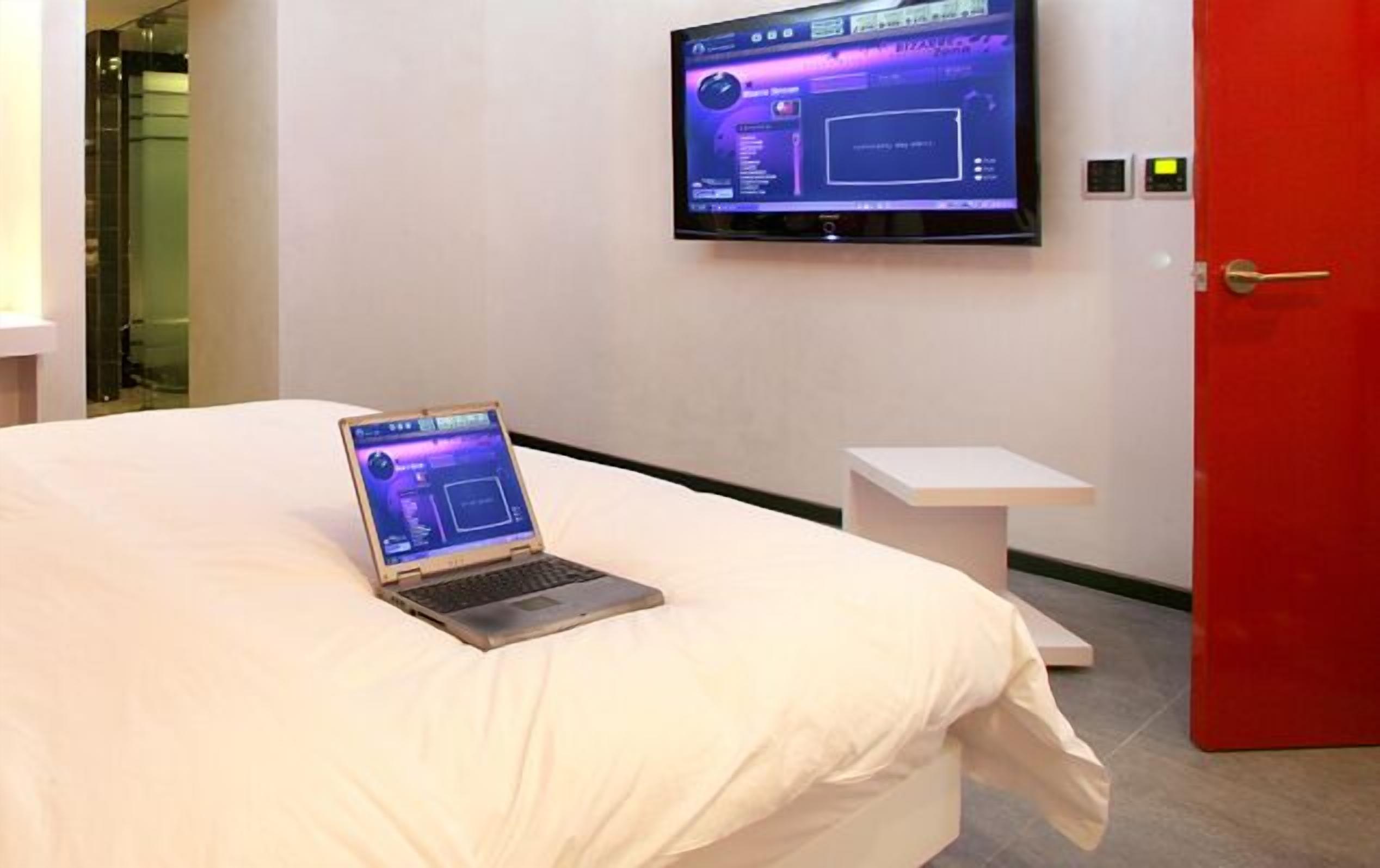 A hotel room with a bed, a laptop on the bed, a TV on the wall, and a red door.