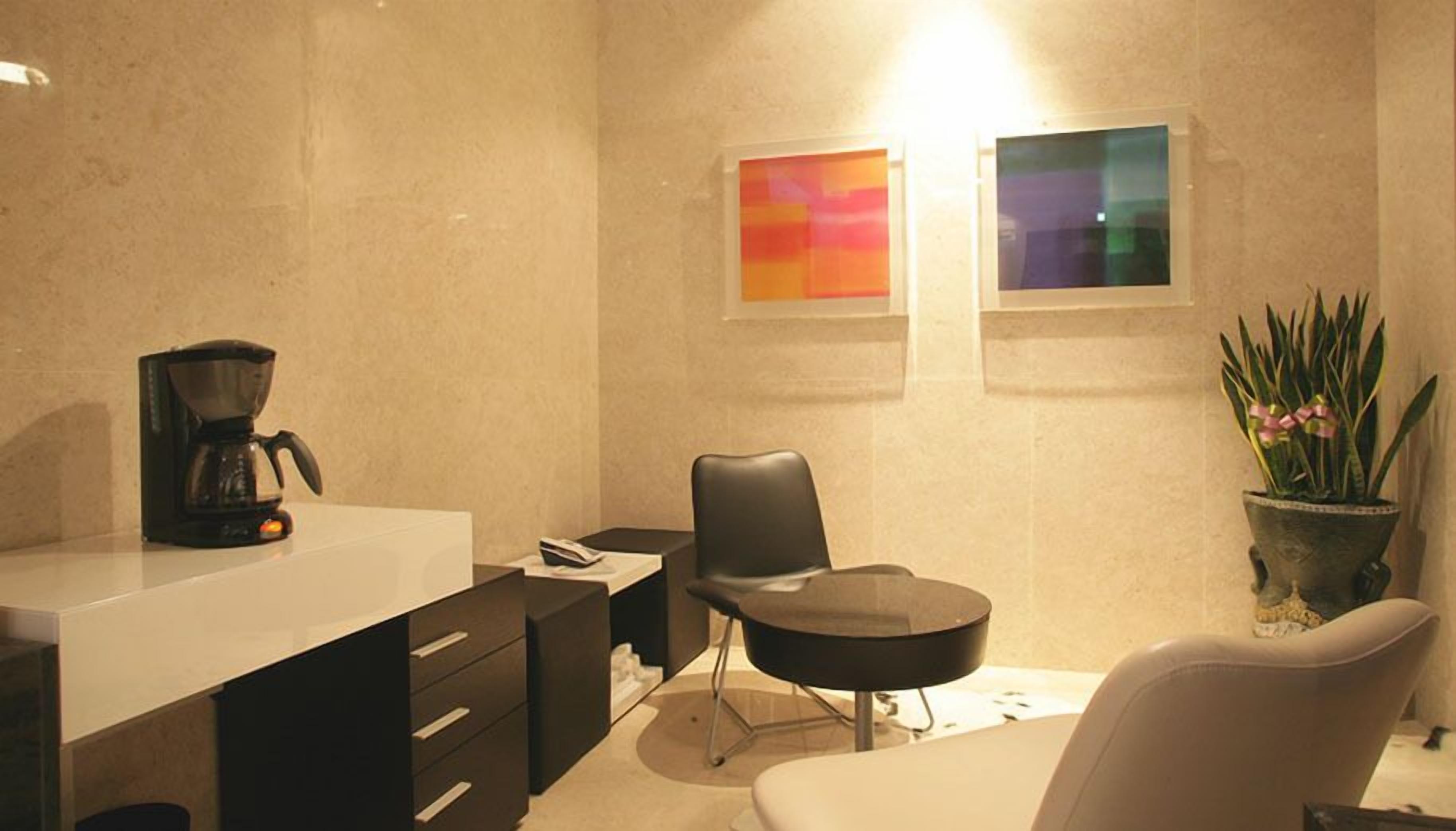 Quarto moderno com escrivaninha branca, cadeira preta, cafeteira, planta em vaso e duas obras de arte emolduradas na parede.