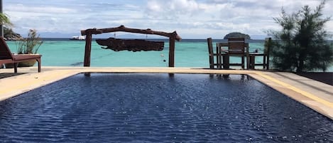 A rectangular swimming pool with a wooden deck and lounge chairs, overlooking a clear blue sea and distant islands.