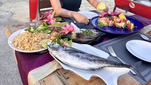 Une table à manger dressée avec une variété de plats, notamment un poisson, une salade et du riz frit, le tout mis en valeur par un décor de plage avec des bateaux et un ciel dégagé.
