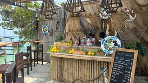 Un bar situé en bord de mer, doté d’une structure en bambou, d’un comptoir en bois et d’un tableau affichant le menu.