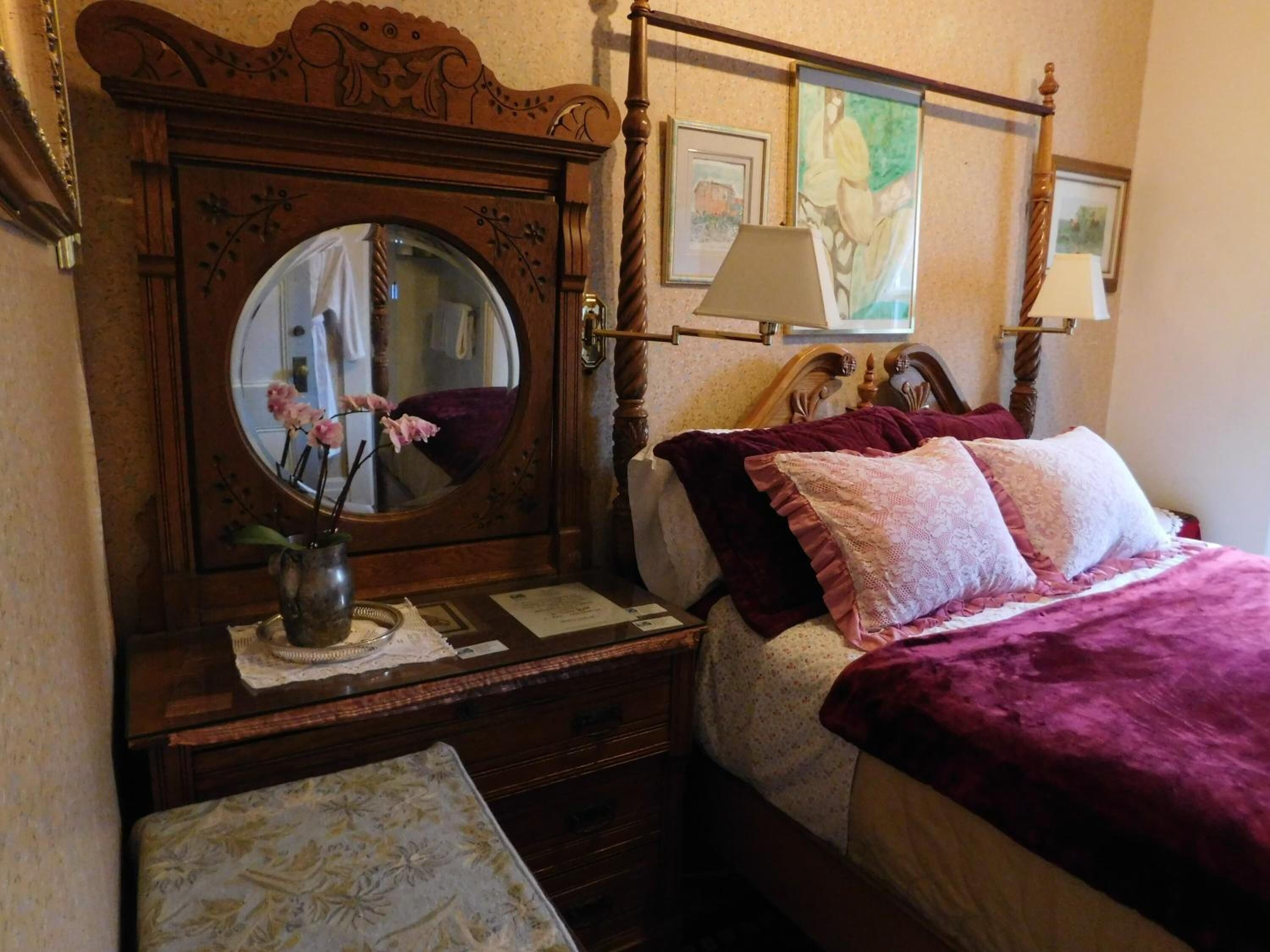 A bedroom with a wooden headboard, a round mirror, a bedside table with a lamp, and a bed with a purple blanket and pillows.