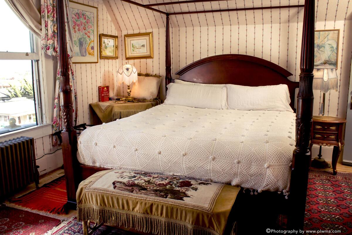 A four-poster bed with a white quilt, a wooden headboard, and a floral-patterned bed runner. There is a nightstand with a lamp and a framed picture on the wall.