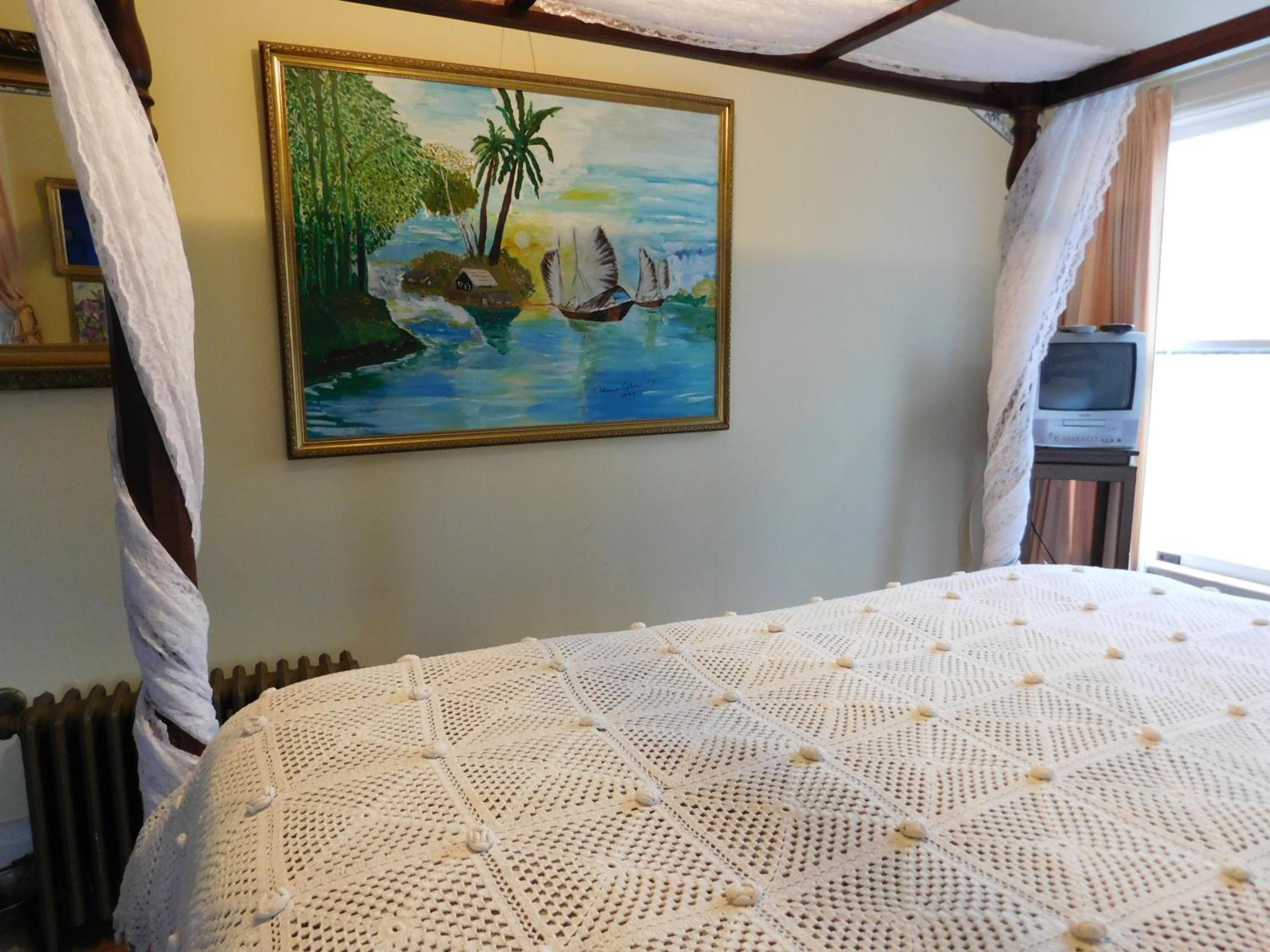 Um quarto com uma cama coberta por um espaleira de malha branca, um quadro de uma cena tropical na parede e uma televisão sobre um suporte à direita.