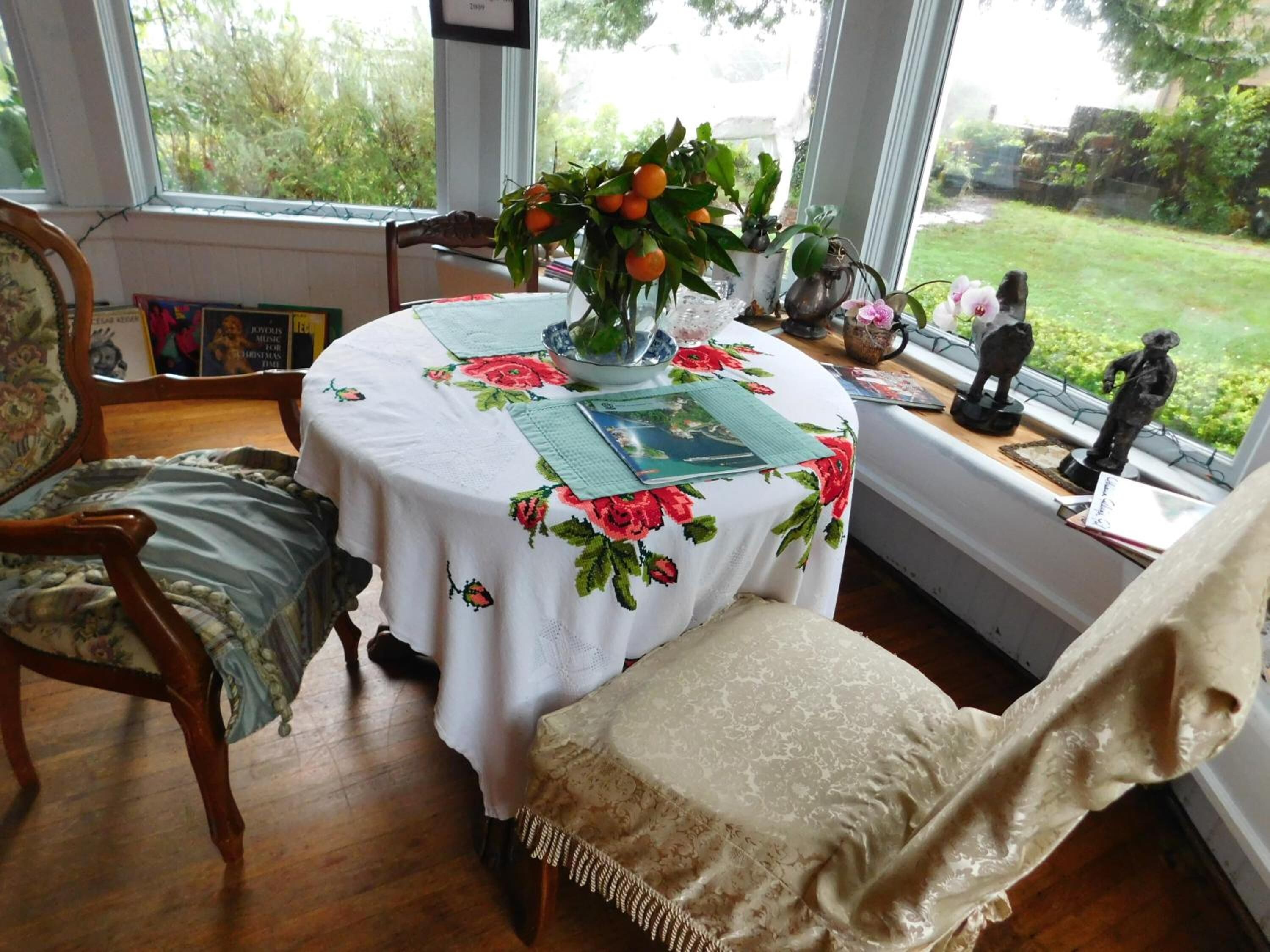 A cozy room with a round table covered by a floral tablecloth, a chair with a cushion, and a window with a view of greenery.