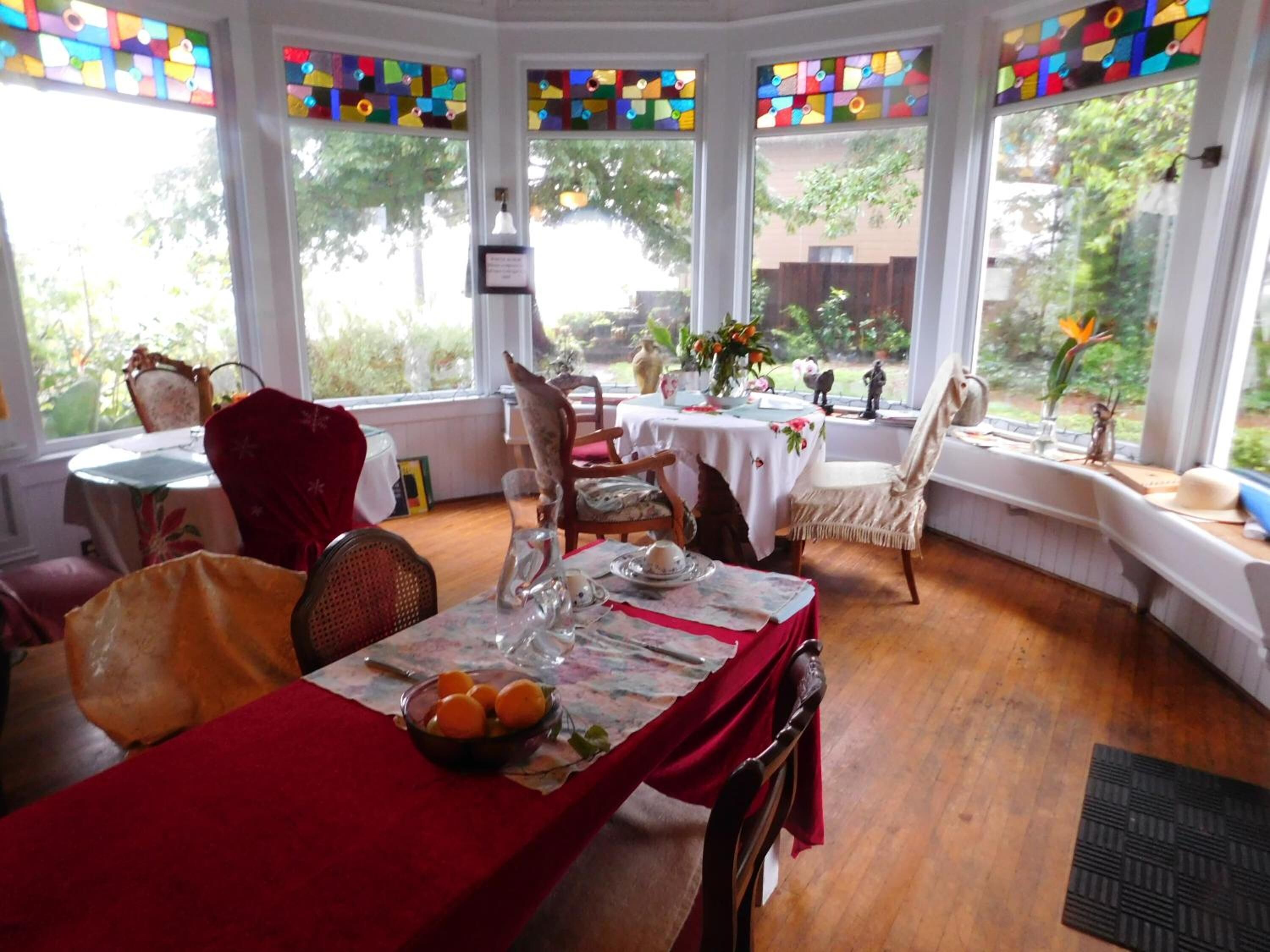 Un comedor con una mesa dispuesta para cuatro personas, sillas alrededor y ventanas amplias con vidrieras de colores.