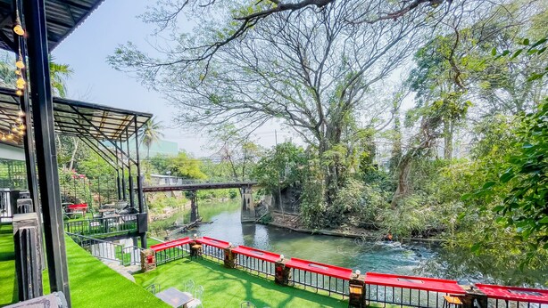 Eine Uferterrasse mit Tischen und Stühlen, eine Brücke und üppiges Grün.