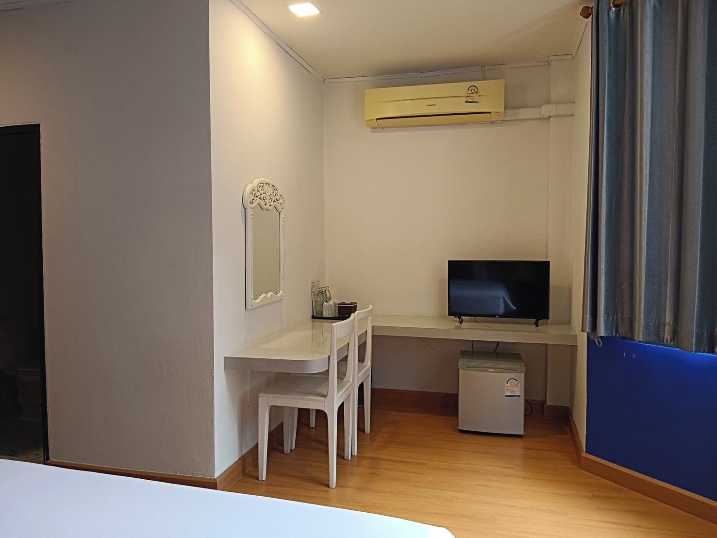 Una habitación compacta con escritorio blanco, televisor, refrigerador pequeño y espejo en la pared.