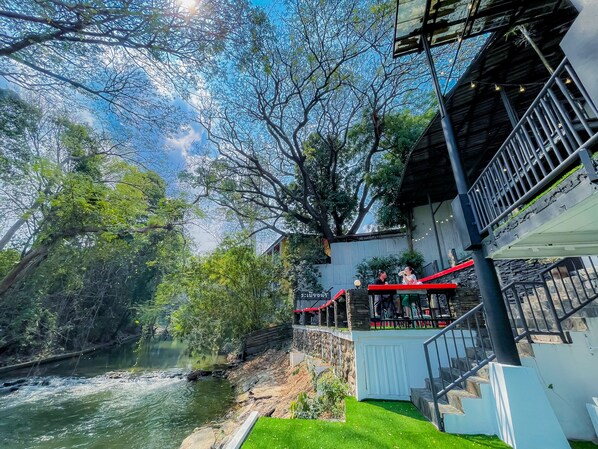 A riverside view with a stone bridge, a wooden deck with red railings, and a building with a white facade. - Rimtarninn Hotel (Pak Chong)