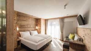 A modern bedroom with a wooden headboard, a bed with white linens, a bedside table with a lamp, and a TV mounted on the wall. - Hotel Pider (La Valle)