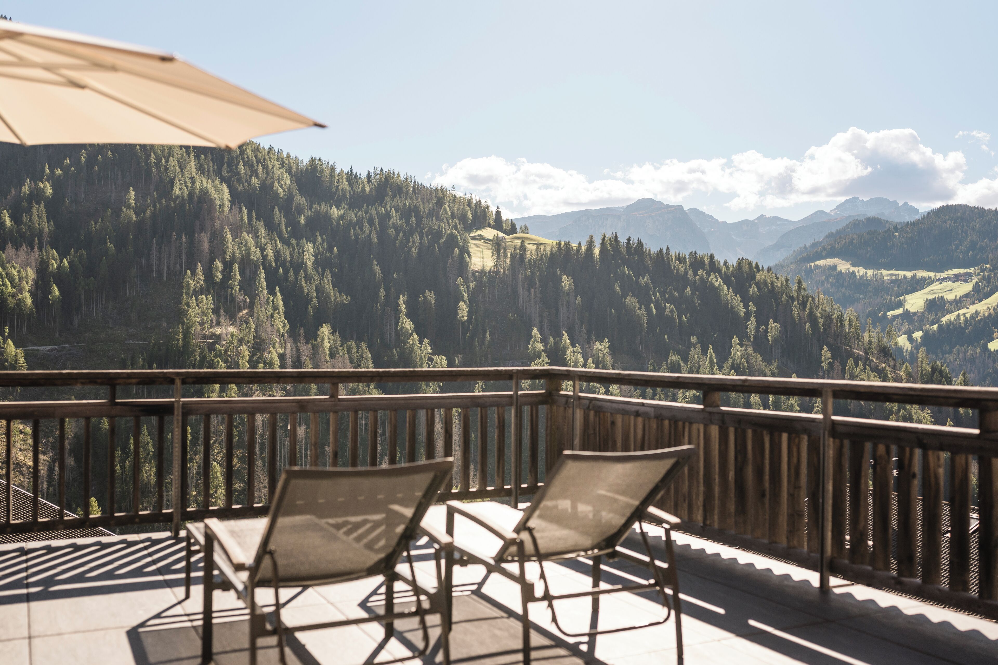A balcony with two lounge chairs and a large umbrella, overlooking a mountainous landscape with dense forests and distant hills.