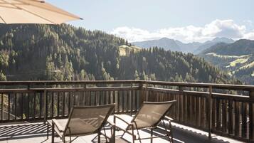 A balcony with two lounge chairs and a large umbrella, overlooking a mountainous landscape with dense forests and distant hills.