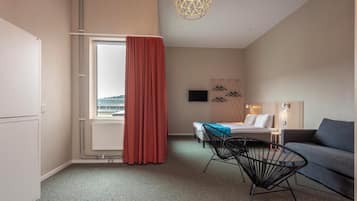 A hotel room with a bed, a desk with a chair, a sofa, a window with curtains, a wall-mounted TV, and a decorative wall piece.
