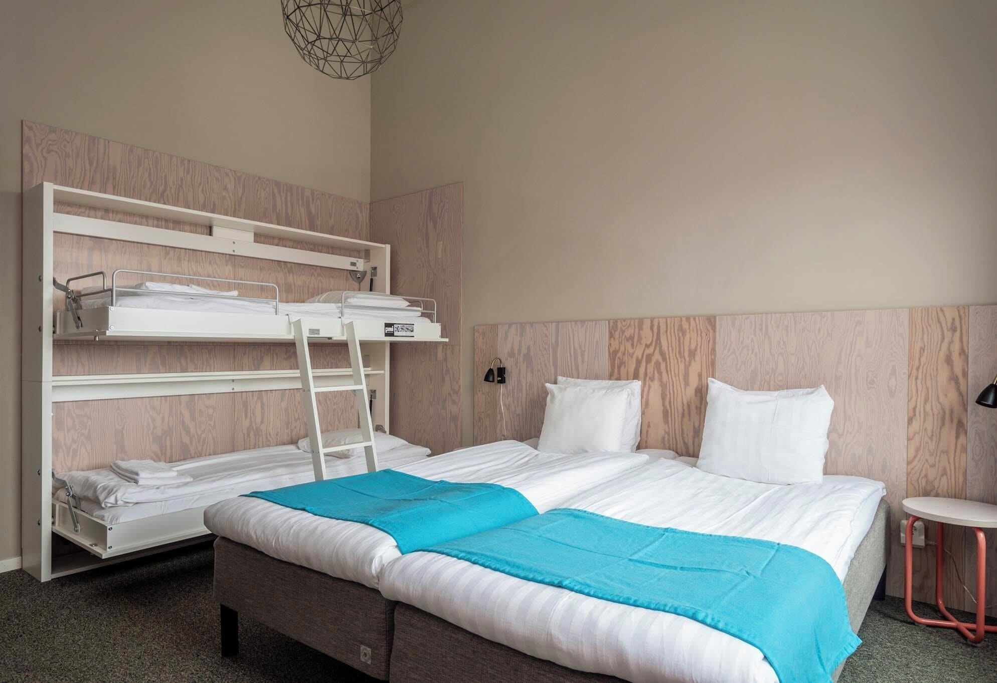 A hotel room with a bunk bed, a single bed, a small table, and a wall-mounted shelf.