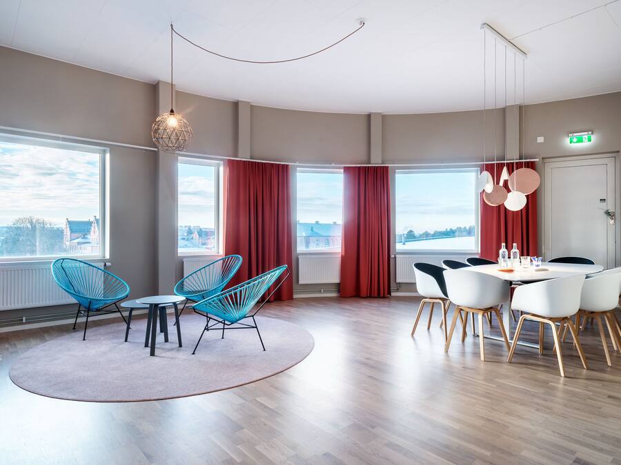 A modern hotel room with large windows, a dining area with white chairs, and a view of the cityscape.