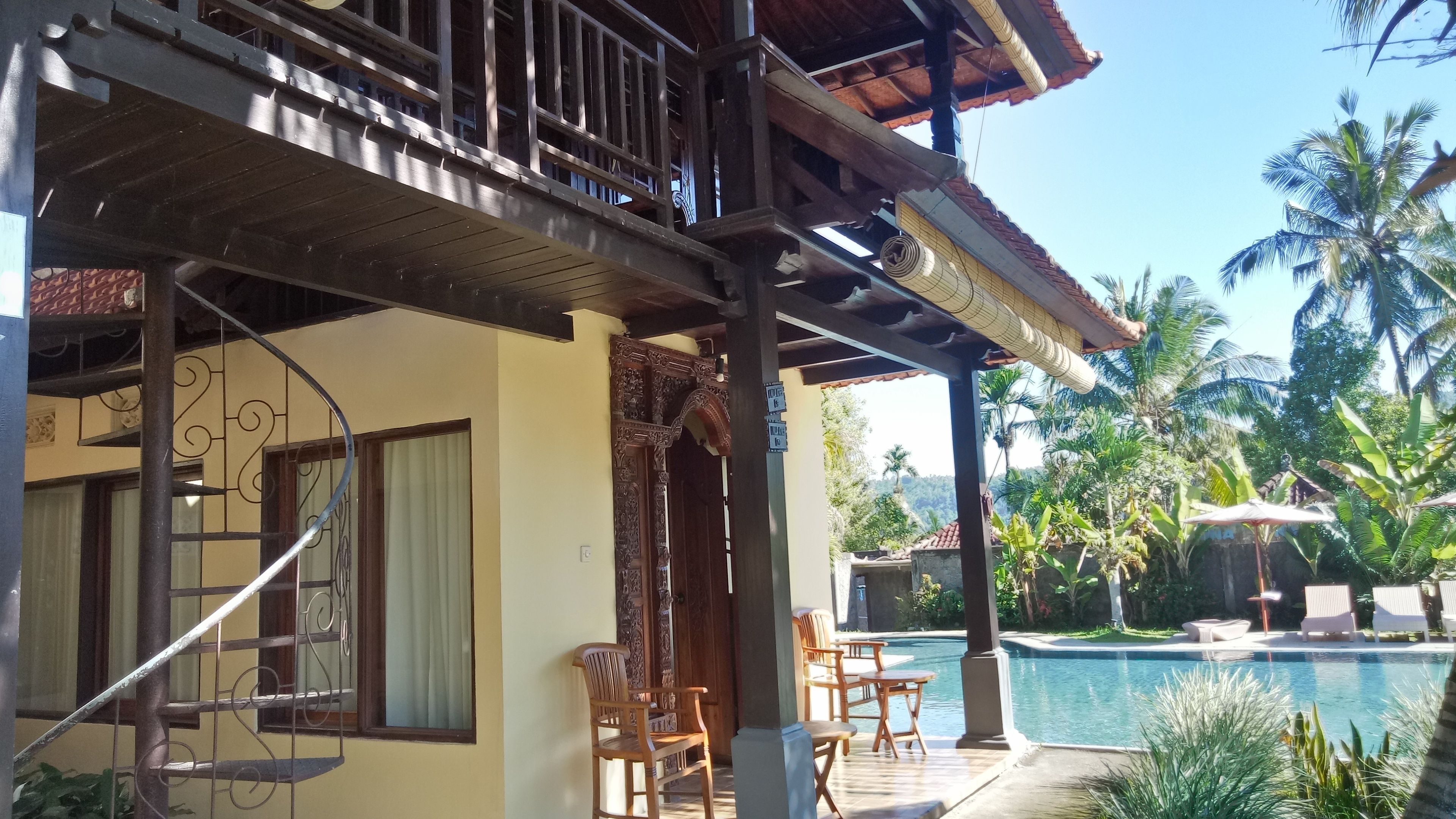 A two-story building with a balcony, a pool area with lounge chairs, and tropical vegetation.