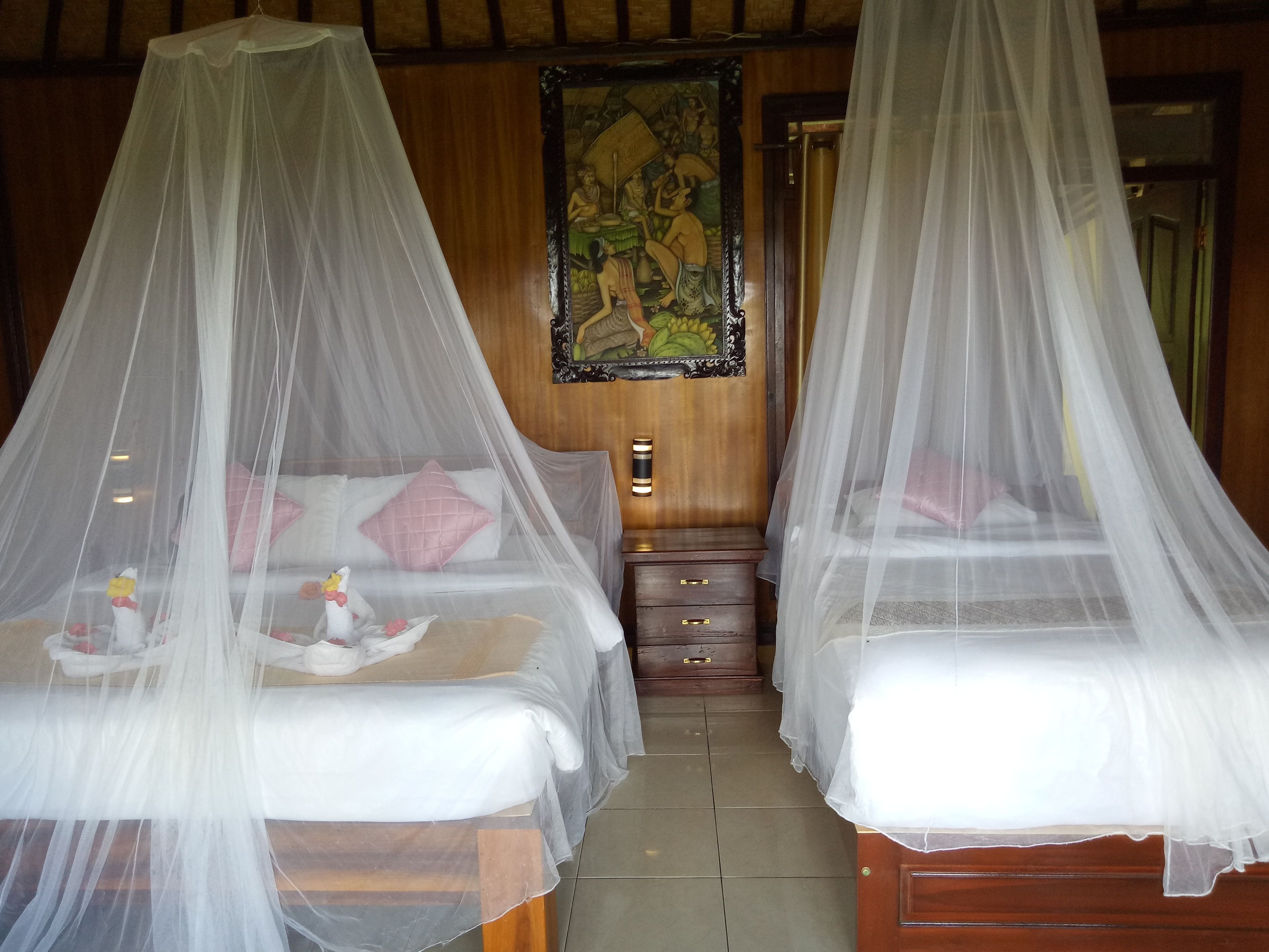 Two beds with white bedsheets and pink pillows, each with a canopy. There is a wooden nightstand between the beds.