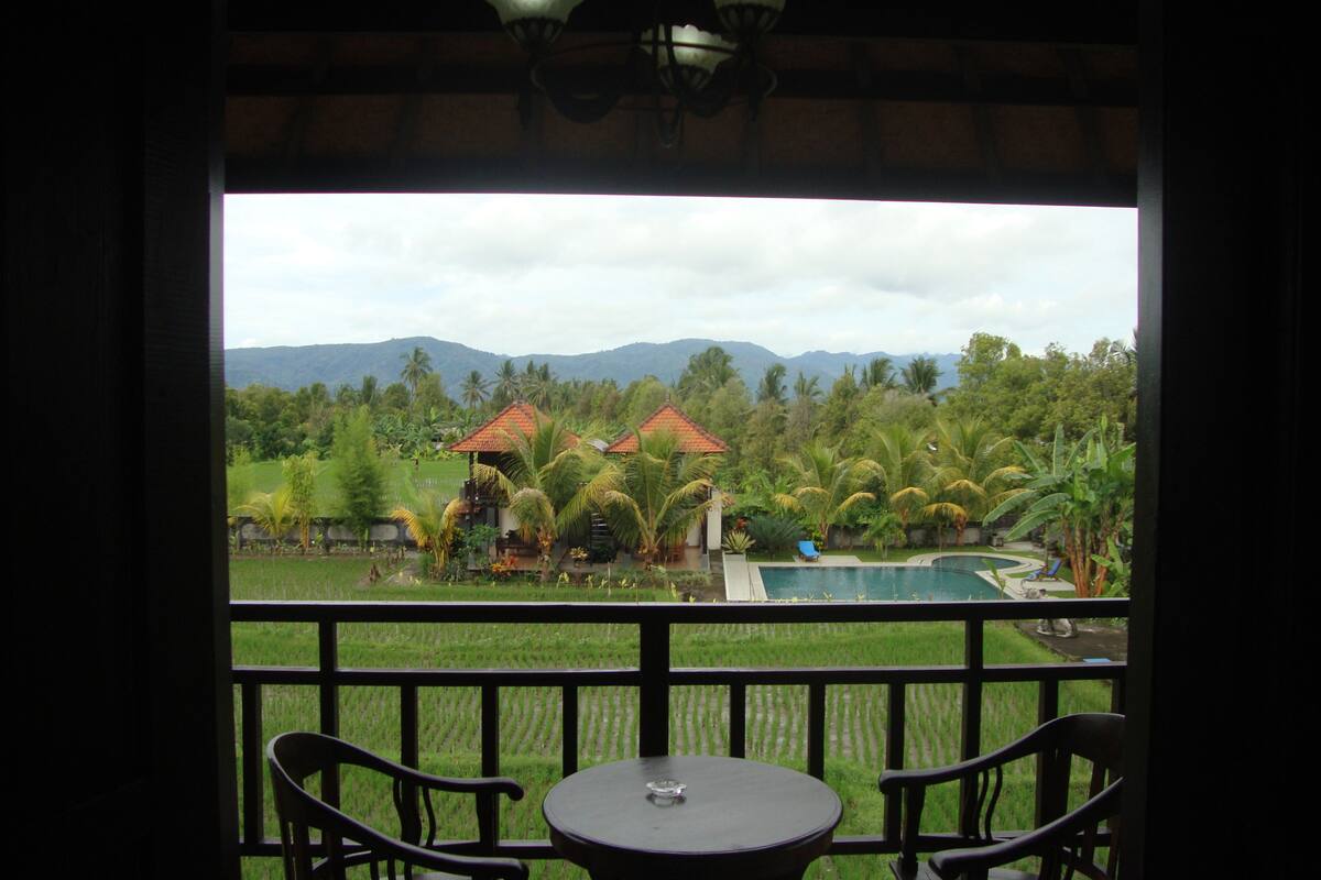 A balcony with a table and chairs, overlooking a pool and lush greenery with palm trees and a mountain range in the background.