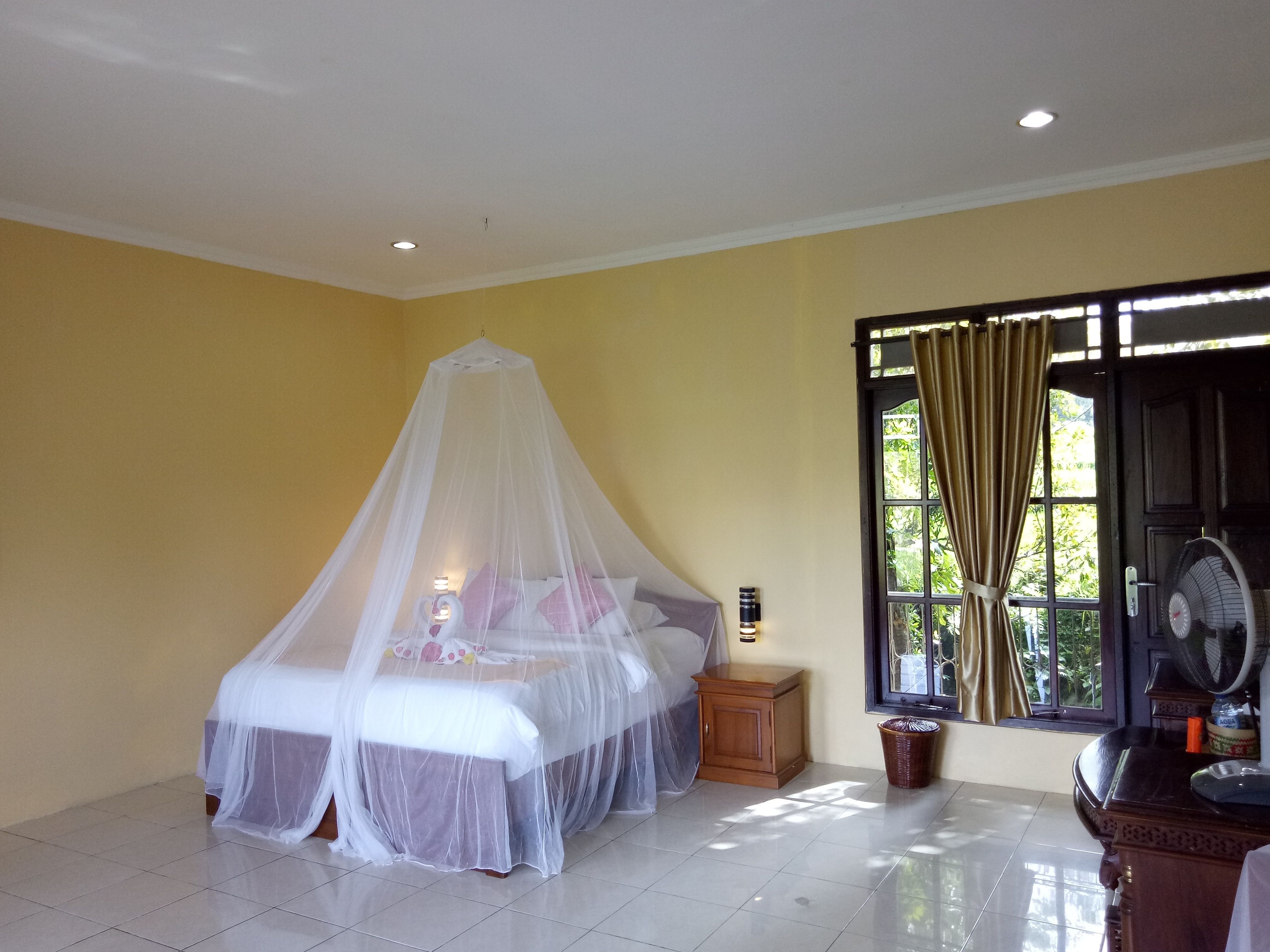 A bedroom with a bed under a mosquito net, a bedside table with a lamp, a window with curtains, and a fan on a wooden table.