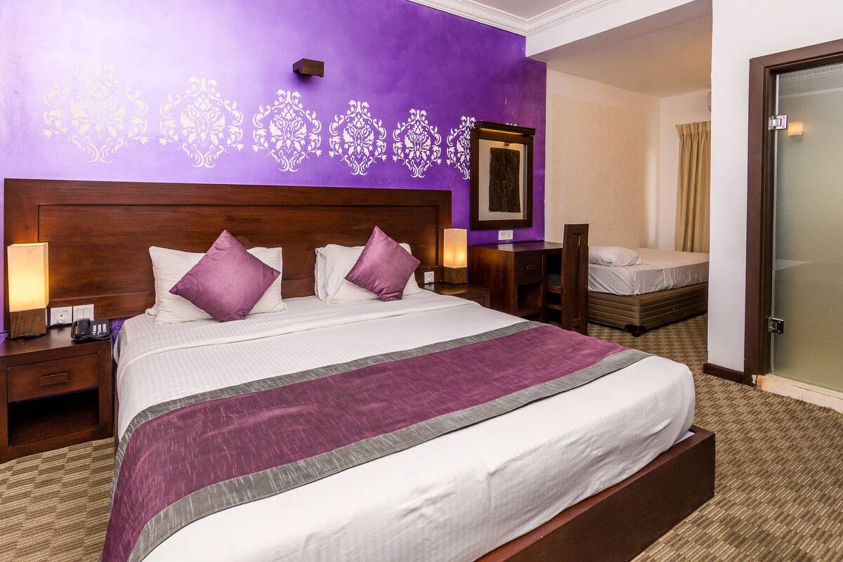 Quarto de hotel com uma cama grande, duas mesas de cabeceira com luminárias, cabeceira de madeira e parede roxa com desenhos decorativos.
