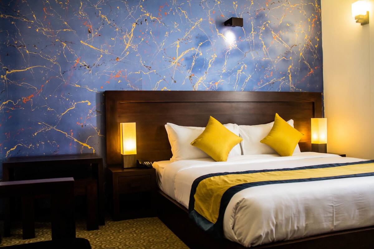 Quarto de hotel com uma cama grande, duas luminárias de cabeceira, um criado-mudo e um banco.