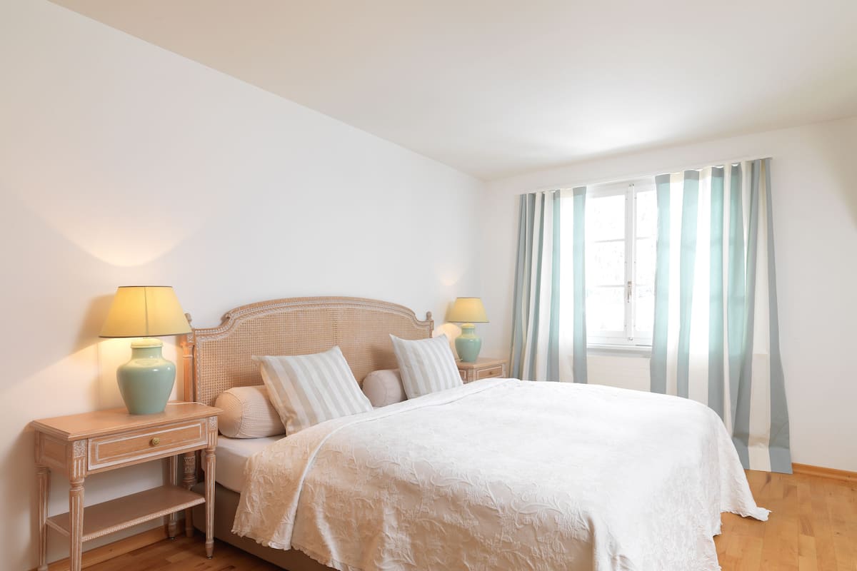 A bedroom with a bed, two bedside tables with lamps, a window with curtains, and a wooden floor.