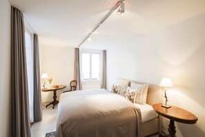 Ein Hotelzimmer mit einem großen Bett, einem Nachttisch mit Lampe, einem kleinen Tisch mit Blumen und einem Fenster mit Vorhängen.