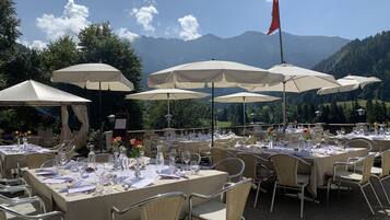 Un área de comedor al aire libre con mesas dispuestas para comensales, rodeada de sombrillas grandes y con vistas a un paisaje montañoso.