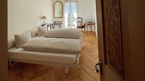 Habitació amb un llit, una cadira, una taula petita i una porta amb un número.