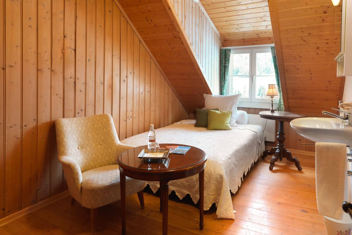 A cozy attic bedroom with wooden walls, a bed with pillows, a chair, a small table, and a bathroom with a sink and mirror.