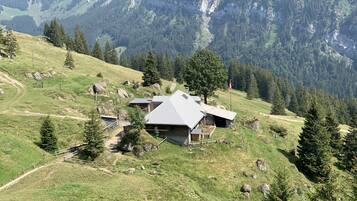 Una cabaña de montaña con techo inclinado, rodeada por un paisaje boscoso y terreno rocoso.