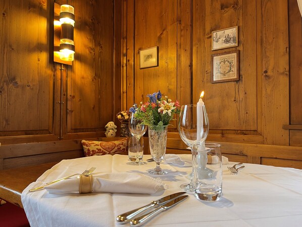 Un salón con paneles de madera y una mesa de comedor dispuesta para dos personas, con una vela, copas de vino y un florero.
