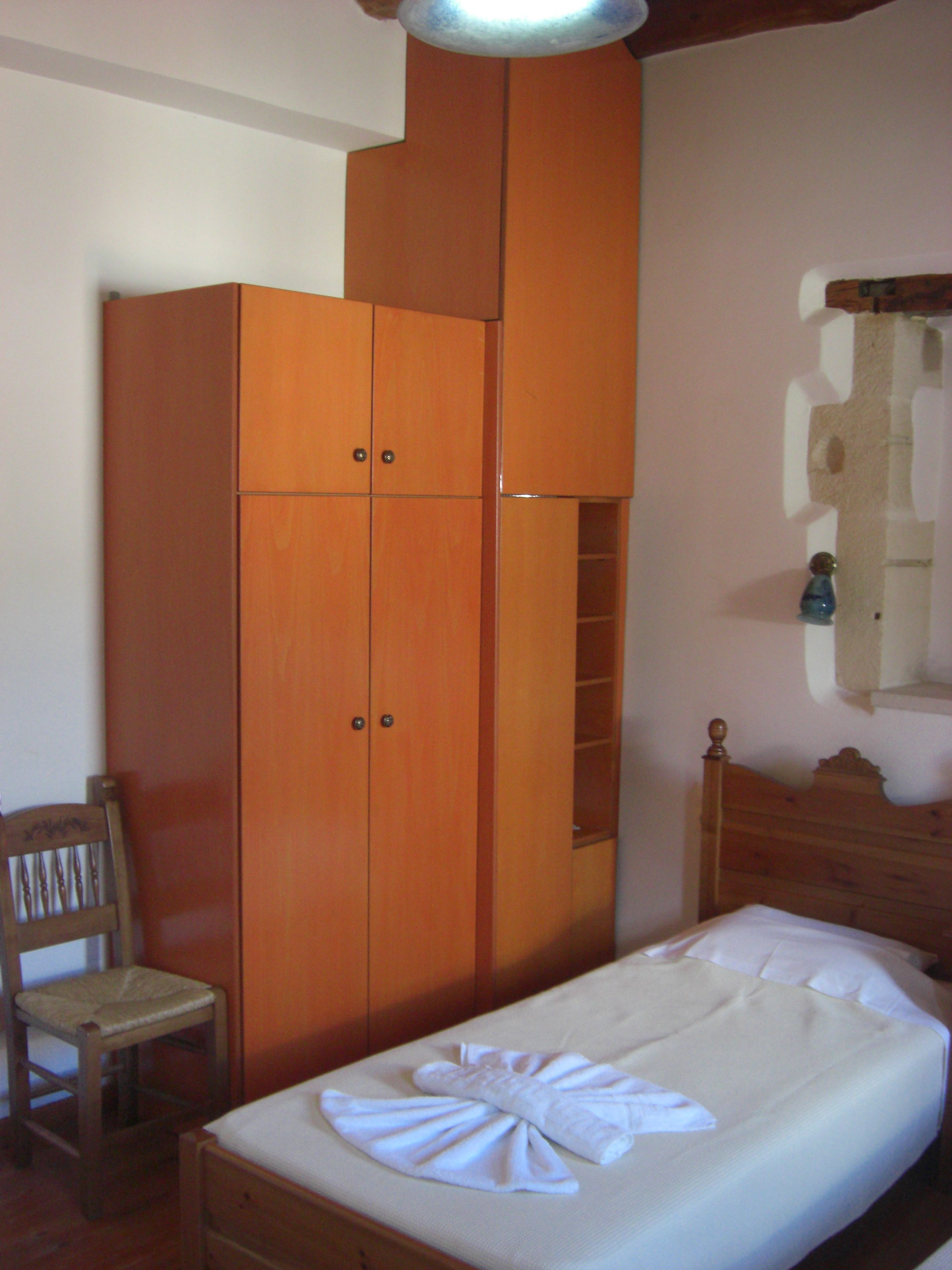 Une chambre avec une armoire en bois, une chaise en bois, un lit recouvert d’une courtepointe blanche et une tête de lit en bois.