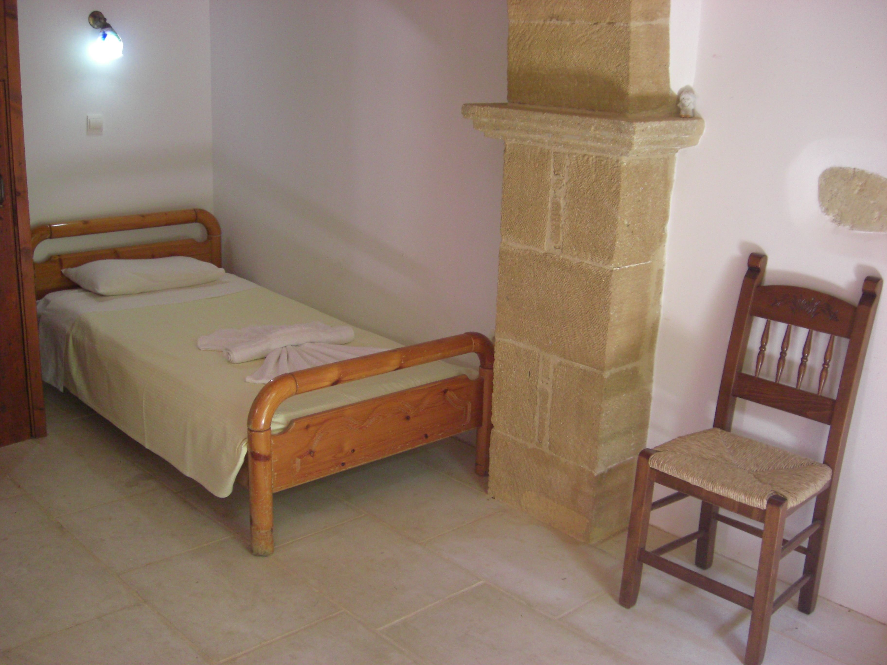 Una habitación pequeña con una cama individual, estructura de madera, un pilar de piedra y una silla de madera.