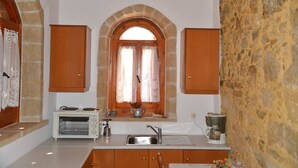 A kitchen with a stone wall, wooden cabinets, a built-in oven, and a window with lace curtains. - Patriko Traditional Residence (Apokoronas)