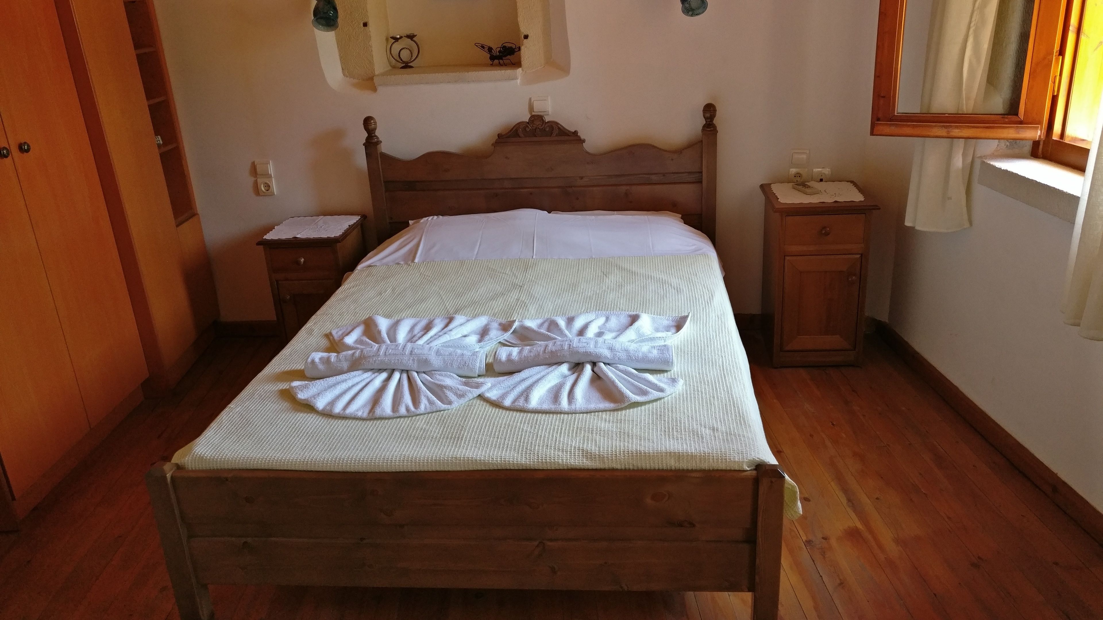 Un lit en bois recouvert d’une courtepointe blanche, avec deux serviettes pliées posées dessus. Une table de chevet en bois se trouve à côté du lit, avec une lampe et une prise électrique sur le mur.