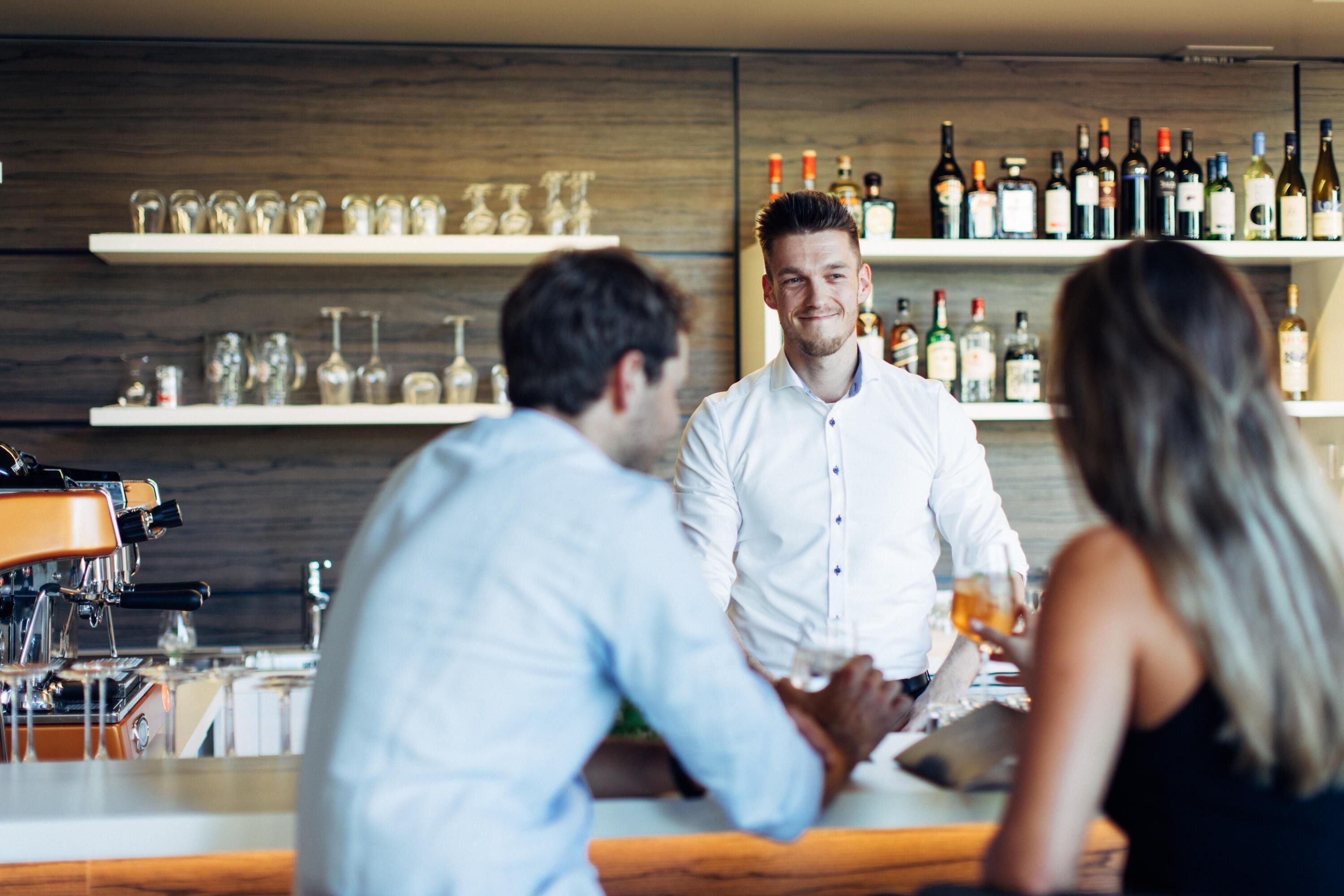 A bar area with shelves of bottles and glasses, a coffee machine, and a counter where people are seated.