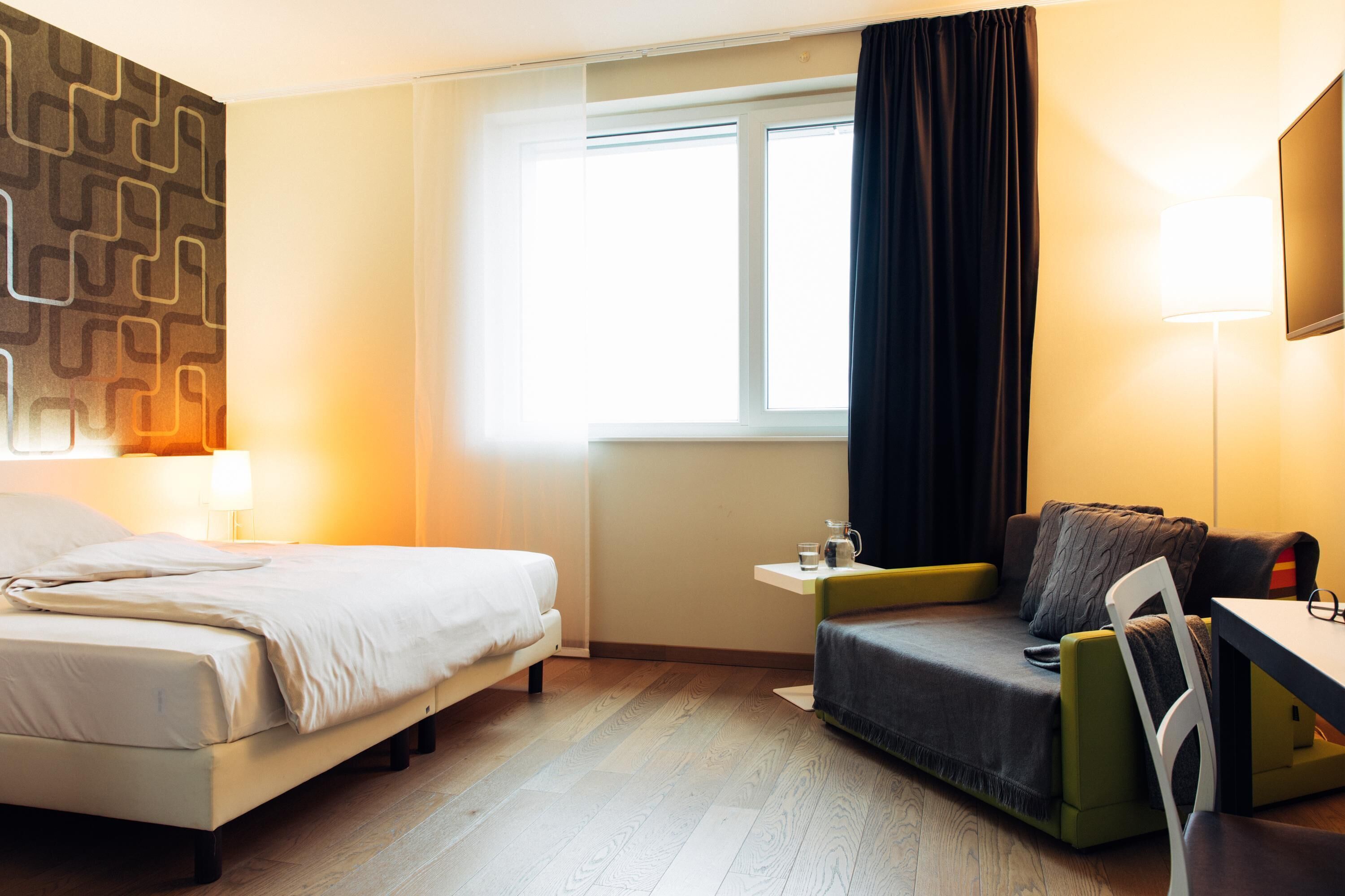 A hotel room with a bed, a desk, a chair, a small table, a window with curtains, and a wall with a geometric pattern.