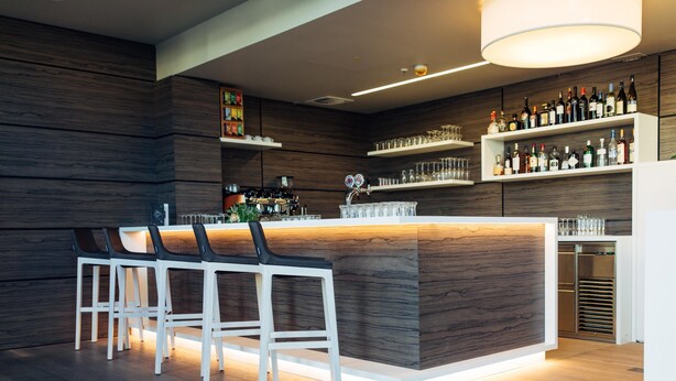A modern bar area with a wooden bar counter, white bar stools, and shelves stocked with bottles and glasses. - harry’s home München Moosach hotel & apartments (Munich)