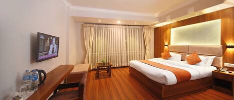 A hotel room with a wooden bed, a TV on the wall, a desk with a chair, and a small table with a plant.